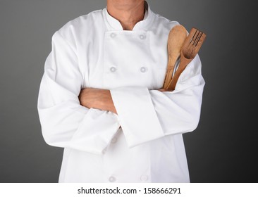 A Chef With His Arms Folded Holding Wood Utensils. Man Is Unrecognizable. Horizontal Format On A Light To Dark Gray Background.