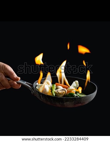 Chef holding a pan with vegetables set ablaze suggesting cooking technic involving sprinkling a liquor over ingredients and briefly igniting fire on them
