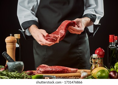 Chef holding a piece of beef meat. Vegetables, bottles with condiments and oil and other ingredients on table. Cooking beef steak. - Shutterstock ID 1731476866