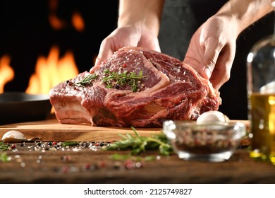 Chef Holding Raw Seasoned Rib Eye Steak Meat Beef On Wooden Chopping Board On A Wooden Table Prepared For Cooking With Flames In The Background. 