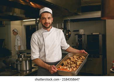 Chef holding tray with baked vegetables while standing in kitchen of restaurant and looking camera - Powered by Shutterstock