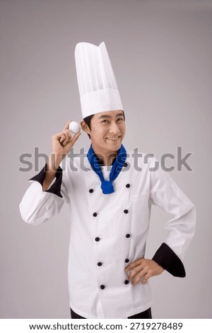 Chef holds an egg and smiles while wearing a hat and white uniform in a simple background