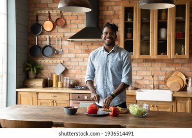 Chef of home kitchen. Portrait of smiling millennial black male hipster enjoy easy cooking food at modern comfortable interior. Happy young afro american guy look at camera distracted of cutting salad - Powered by Shutterstock