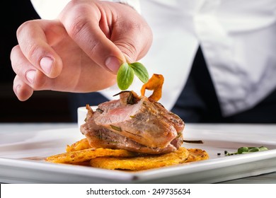 Chef in hotel or restaurant kitchen cooking, only hands. Prepared meat steak with potato or celery pancakes.He is working on the herb decoration.