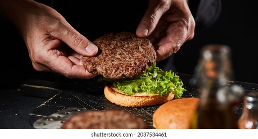 Chef Making A Burger With A Succulent Homemade Grilled Beef Patty Placing It On Top Of Fresh Lettuce On A Toasted Bun In Close Up On His Hands