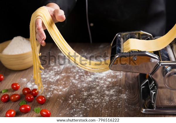 Chef making spaghetti noodles\
with pasta machine on kitchen table with some ingredients\
around.\
