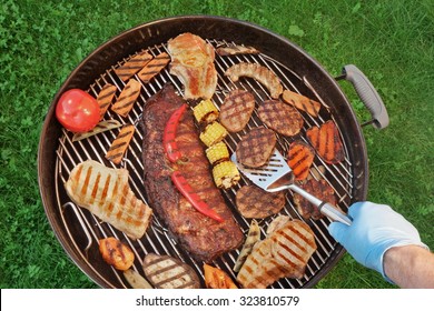 Chef Male Hand With Spatula And BBQ Grill With Roasted Different Kind Of Meat And Vegetables, Overhead View, Backyard Party Or Picnic Concept