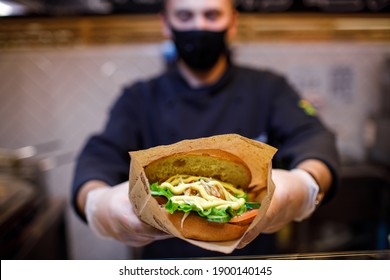 A Chef In A Mask And Gloves Holds Out A Burger Wrapped In Parchment Paper