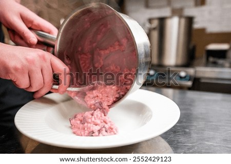 A chef plating a vibrant pink risotto from a stainless steel pot onto a white plate in a professional kitchen. Steam rises, creating an inviting atmosphere around the dish.