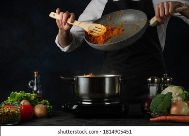 The chef pours chopped tomatoes into the pan from the frying pan by the kitchen spatula. Chef black apron cooking tasty dish isolated on dark background. Restaurant's meal. Photo for culinary. - Shutterstock ID 1814905451