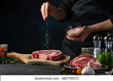 Chef pours sea salt on raw steak on wooden chopped board. Backstage of preparing grilled pork meat at restaurant kitchen on dark blue background. Frozen motion. Cooking process.