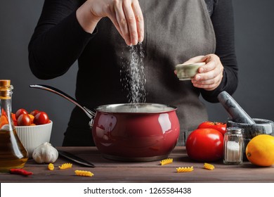The chef preparations spaghetti and pasta, salt water, against a dark background, the concept of cooking. Woman salting water before cooking pasta fusilli - Shutterstock ID 1265584438
