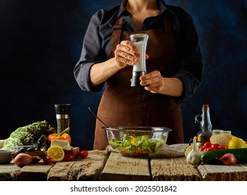 The Chef Prepares A Classic Greek Salad. Adds Spices To A Large Glass Salad Bowl. Bright Paint Ingredients. Dark Background, Wood Texture. Step By Step Recipe, Restaurant, Hotel, Fitness Bar.