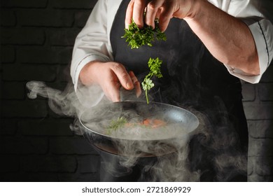 A chef prepares a dish by adding parsley to a hot pan. Steam rises from the pan, showing lively cooking activity in a kitchen. The setting is busy and focused on food.