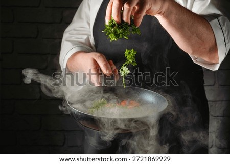 A chef prepares a dish by adding parsley to a hot pan. Steam rises from the pan, showing lively cooking activity in a kitchen. The setting is busy and focused on food.