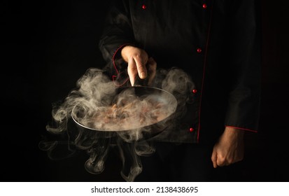 Chef Prepares Food In A Frying Pan With Steam On A Black Background. The Concept Of Restaurant And Hotel Service. Asian Cuisine.