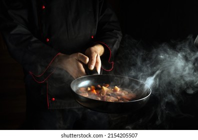 The Chef Prepares Food In A Frying Pan With Steam On A Black Background. The Concept Of Restaurant And Hotel Service. Asian Cuisine