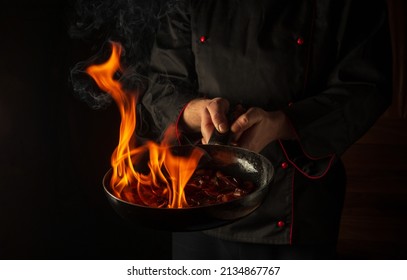 Chef Prepares Vegetable Food In A Frying Pan With A Flame Of Fire. The Concept Of Restaurant And Hotel Service. Flamber Or Cooking Over Natural Fire. Grande Cuisine.