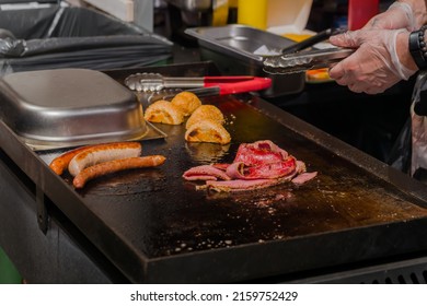 Chef Preparing Burger Buns, Bacon Slices And Long Sausages On Grill At Summer Local Food Market - Close Up View. Outdoor Cooking, Gastronomy, Cookery, Street Food Concept