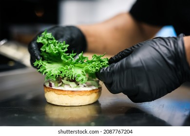 chef preparing a burger by adding fresh  lettuce on burger bread , and wearing black gloves