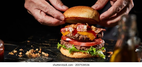 Chef preparing a delicious beef burger with full trimmings including salad ingredients, grilled bacon and cheese placing a toasted bun on top in close up - Powered by Shutterstock