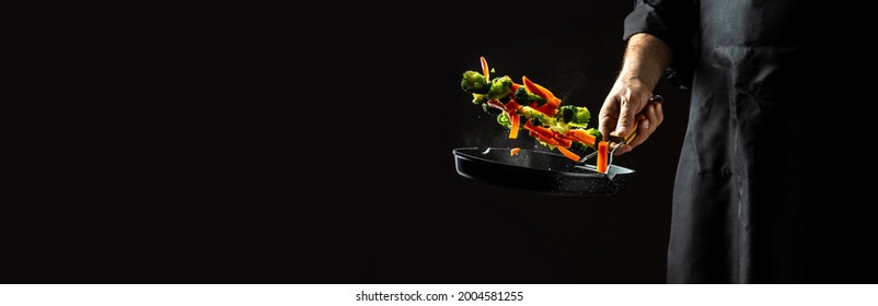 chef preparing food in the kitchen of a restaurant. flying vegetables scattering in a freeze motion of a cloud midair on black. Cook hands with vegetables on a pan.