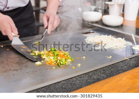 Chef preparing fresh vegetable stir fry over a hot plate turning it with a spatula as he cooks Asian cuisine at a restaurant, close up of his hands