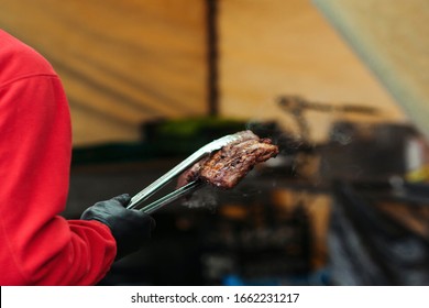 Chef preparing meat on the grill. Street food. Cooking grilled pork ribs. Male hand with tongs holding hot pork ribs. Fresh grilled meat. Street market. Food catering, party. Street festival.