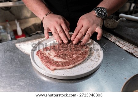 Chef preparing a raw beef steak by pressing seasoning and spices onto the surface on a stainless steel worktop.
