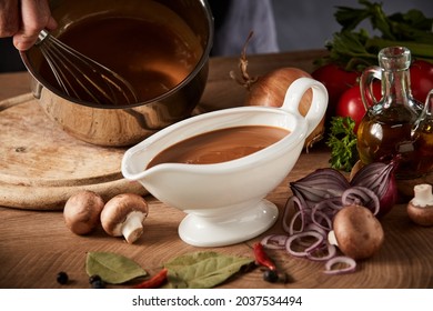 Chef preparing a serving of delicious spicy rich gravy whisking it in a pot with a close up view on a full sauce boat or pitcher in the foreground
