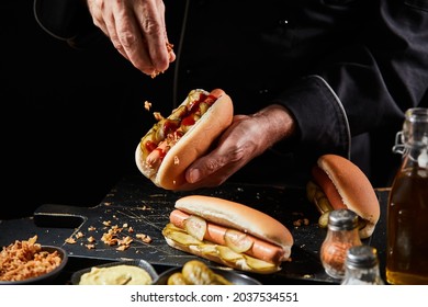 Chef preparing a tasty hot dog with a frankfurter on a fresh roll adding extra trimmings sprinkling crispy bacon bits onto the pickles and sausage