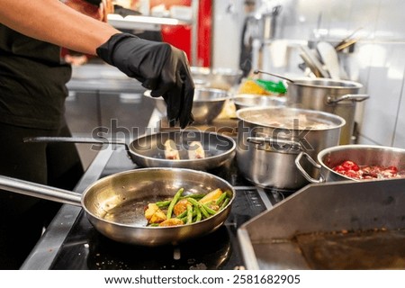 A chef in a professional kitchen prepares a dish, adding green beans and potatoes to a skillet. Various pots and pans are seen on the stove, creating a bustling culinary environment.