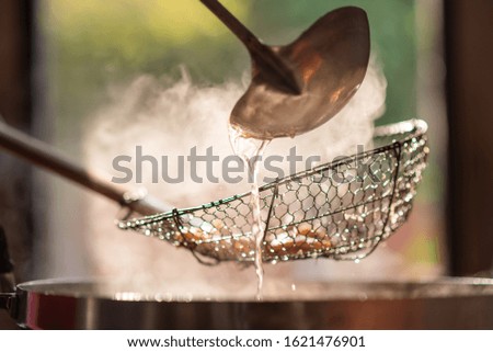 The chef in the restaurant is cooking while using the dipper in a large pot. The water is boiling and the mass of steam reflected in the morning light.