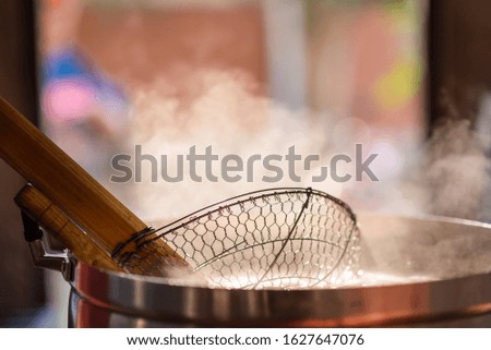 The chef in the restaurant is cooking while using the dipper and sieve for scalding in a large pot. The water is boiling and the mass of steam reflected in the morning light.