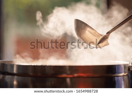 The chef in the restaurant is cooking while using the dipper in a large pot. The water is boiling and the mass of steam reflected in the morning light.