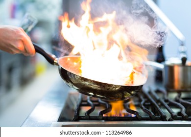 Chef In Restaurant Kitchen At Stove With Pan, Doing Flambe On Food