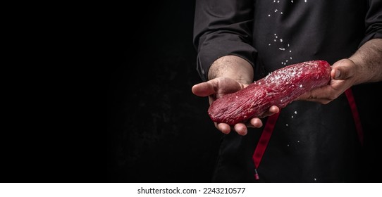 Chef salts steak in a freeze motion with rosemary and spices. Preparing fresh beef or pork on a dark background. Long banner format. - Shutterstock ID 2243210577