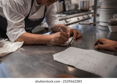 A chef is seen writing notes and planning a menu in a bustling restaurant kitchen. The environment is dynamic, showcasing teamwork and culinary artistry in a professional setting. - Powered by Shutterstock