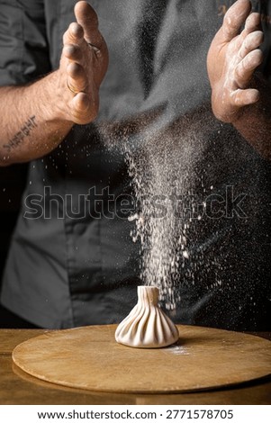 Chef shaping khinkali dumpling while flour falls from hands creating dynamic motion on wooden board in kitchen