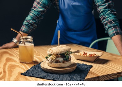 Chef Showing His Vegan Burger With Lettuce And Sauce, Fries With Ketchup And Healthy Drink On A Wooden Table, Vegetarian Food And Gourmet Lifestyle Concept, Selective Focus