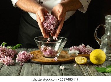 A chef skillfully combines acacia flowers with lemon slices in a clear bowl. Fresh herbs and water accompany the vibrant ingredients on a wooden table, creating a fragrant mixture.
