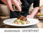 A chef skillfully drizzles olive oil over a beautifully plated dish featuring a seared piece of meat and gnocchi, set in a modern kitchen. Vibrant colors highlight the culinary artistry.