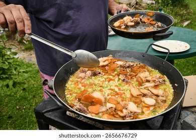 Chef skillfully using a ladle to prepare a vibrant seafood paella in a spacious pan over a portable gas stove in an outdoor setting