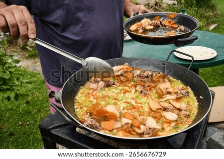 Chef skillfully using a ladle to prepare a vibrant seafood paella in a spacious pan over a portable gas stove in an outdoor setting