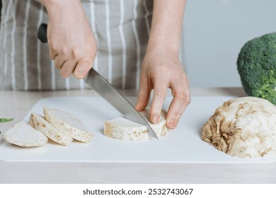 A chef slices celeriac root with knife on a white cutting board, preparing the fresh root vegetable for cooking - Powered by Shutterstock