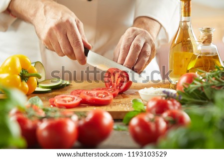 Chef slicing fresh tomatoes for a salad in a low angle view of the knife and chopping board over assorted fresh vegetable ingredients