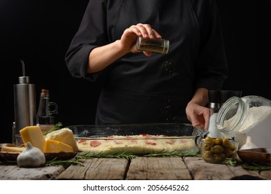 The chef sprinkles the dough prepared for baking with spices. Preparation of traditional Italian focaccia bread. Ingredients. Wooden texture. Dark background. Country style. Restaurant, hotel.