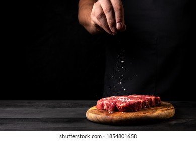 The Chef Sprinkles Salt On A Fresh Steak On A Cutting Board On A Black Background On A Wooden Table. Horizontal Image. With Free Space For Logo Text Labels