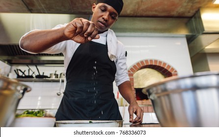 Chef Sprinkling Spices On Dish In Commercial Kitchen. African Male Cook Preparing Food In Restaurant Kitchen, Adding Seasoning.