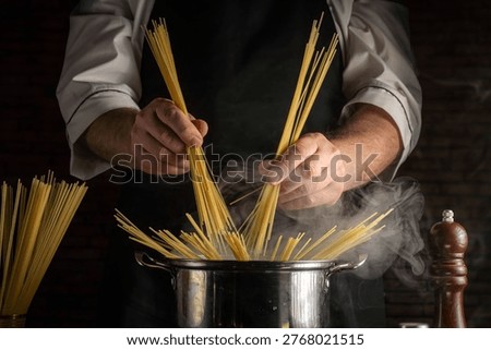 A chef stands in a kitchen, adding bundles of raw spaghetti into a pot of boiling water. Steam rises from the pot. Ingredients for pasta are spread around the area.